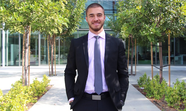 Man in a black suit standing in the middle of an urban outdoor area with trees. He is smiling and resting his hands inside his pocket.