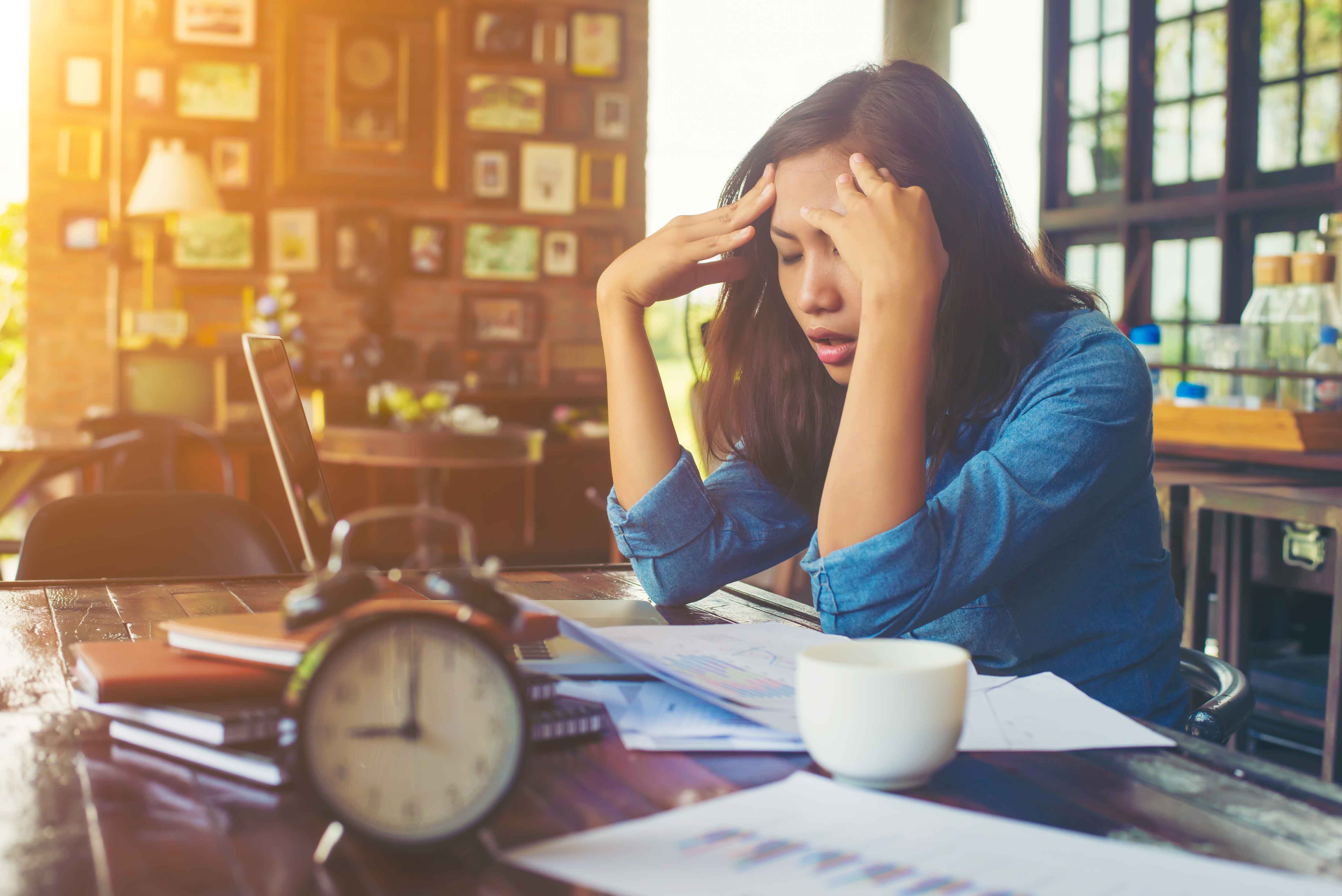 A stressed out woman holding her forehead while sitting at a desk full of papers, a coffee cup and an alarm clock.