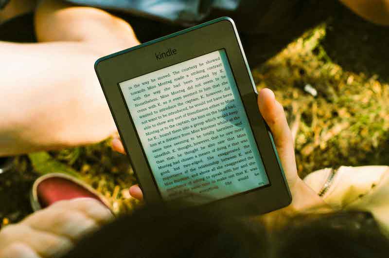 A portable gadget showing a digital version of a book. Point-of-view of a person reading the device while sitting on grass.
