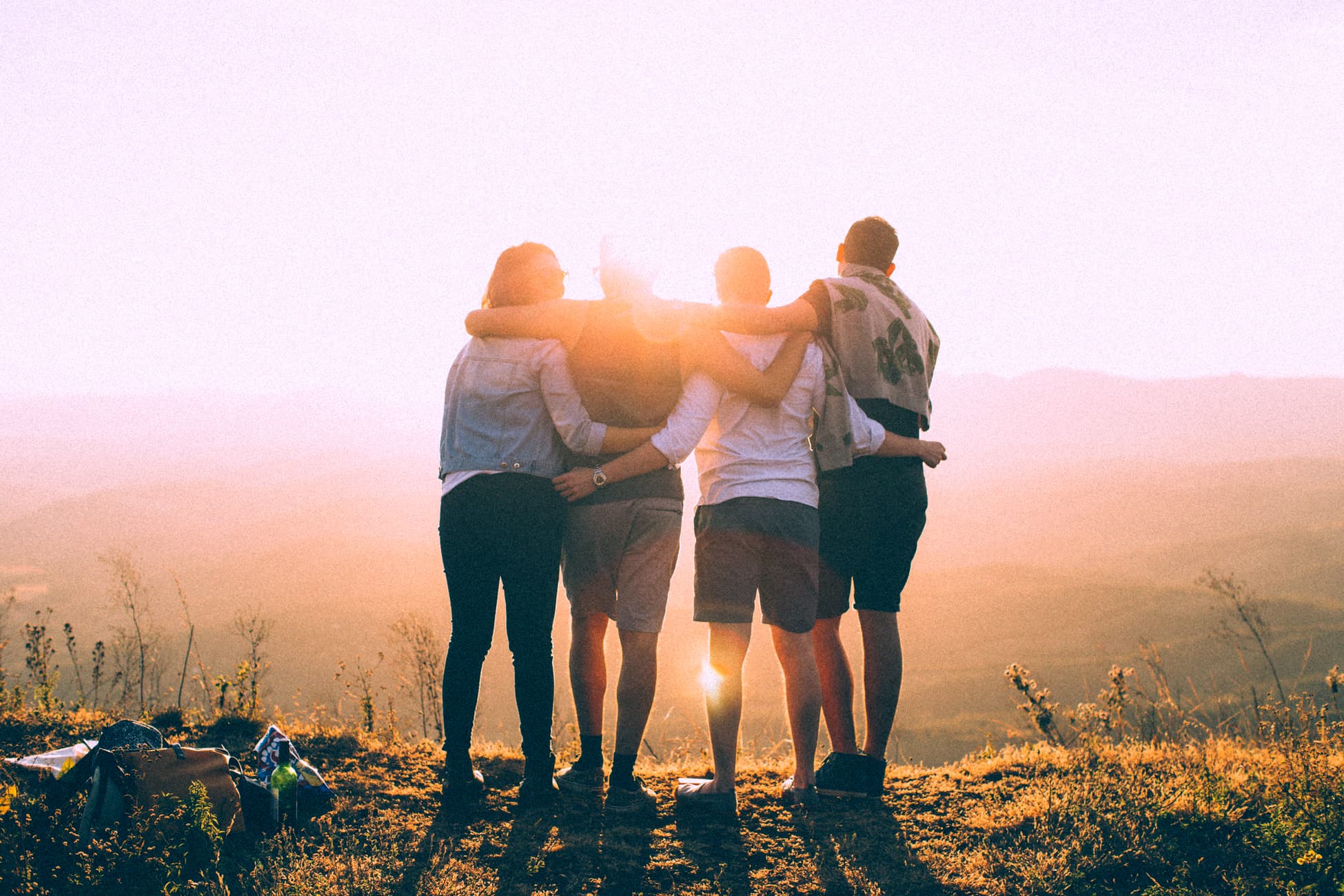 four friends wrap arms around each other. they stand on the edge of a mountain as they gaze off into the sunset