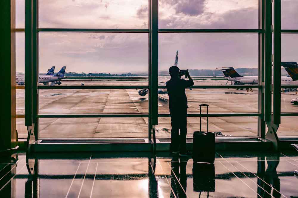 silhouette of man standing beside his luggage in front of huge airport window. he takes a photo of airplanes parked on the runway