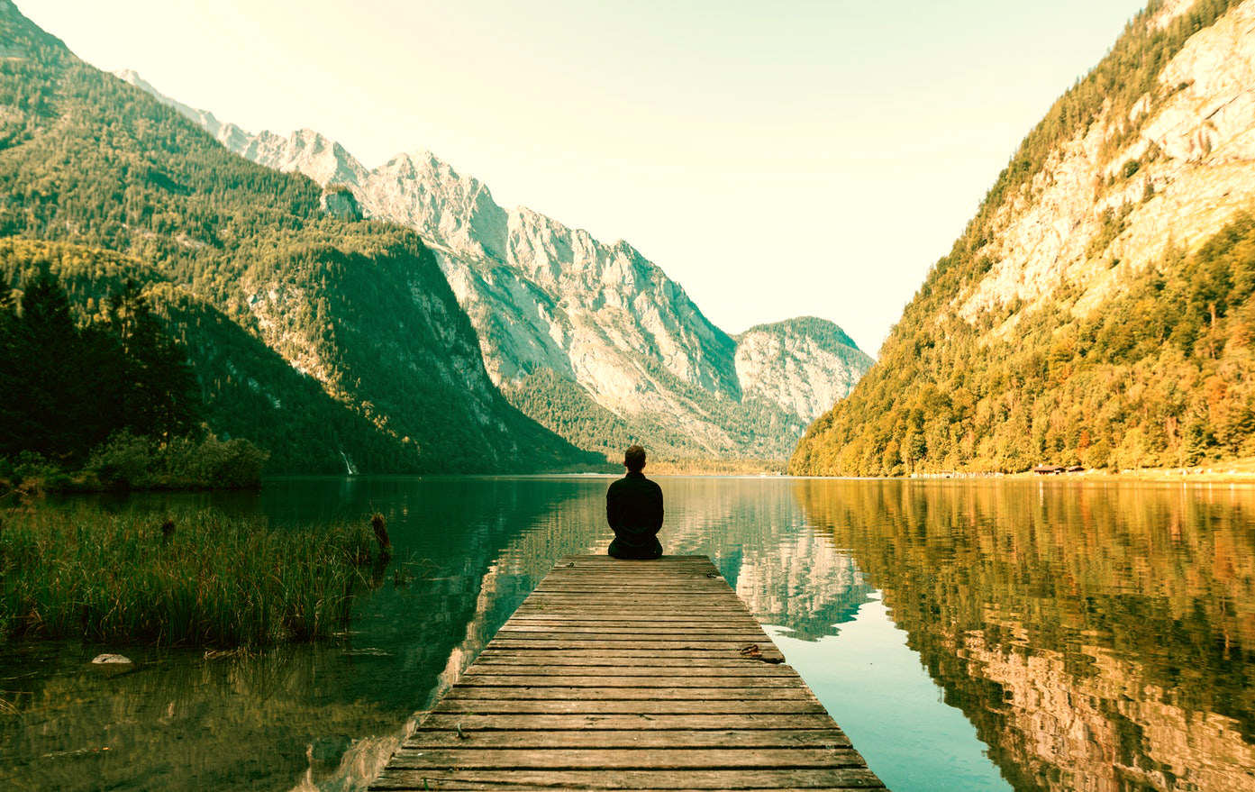 A man sitting on the end of a dock facing a lake and mountains
