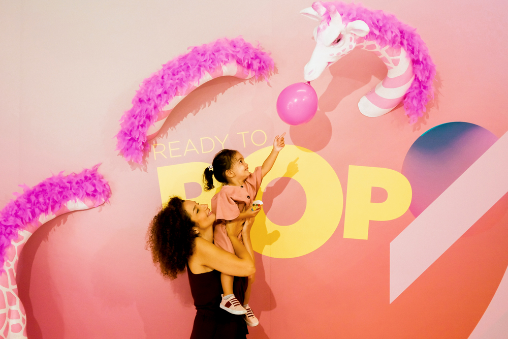 mother lifting up her young daughter as she reaches for a balloon hanging from a pink giraffe's mouth