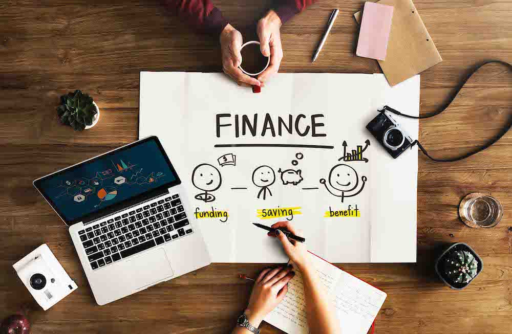 hands gathered around a desk. Holding cup of coffee, writing on paper. "Finance" written on paper