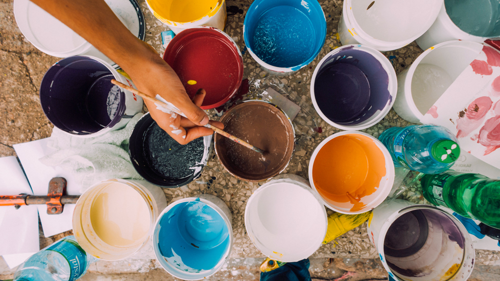 hand dipping a paintbrush into a can of paint amid other jars of paint
