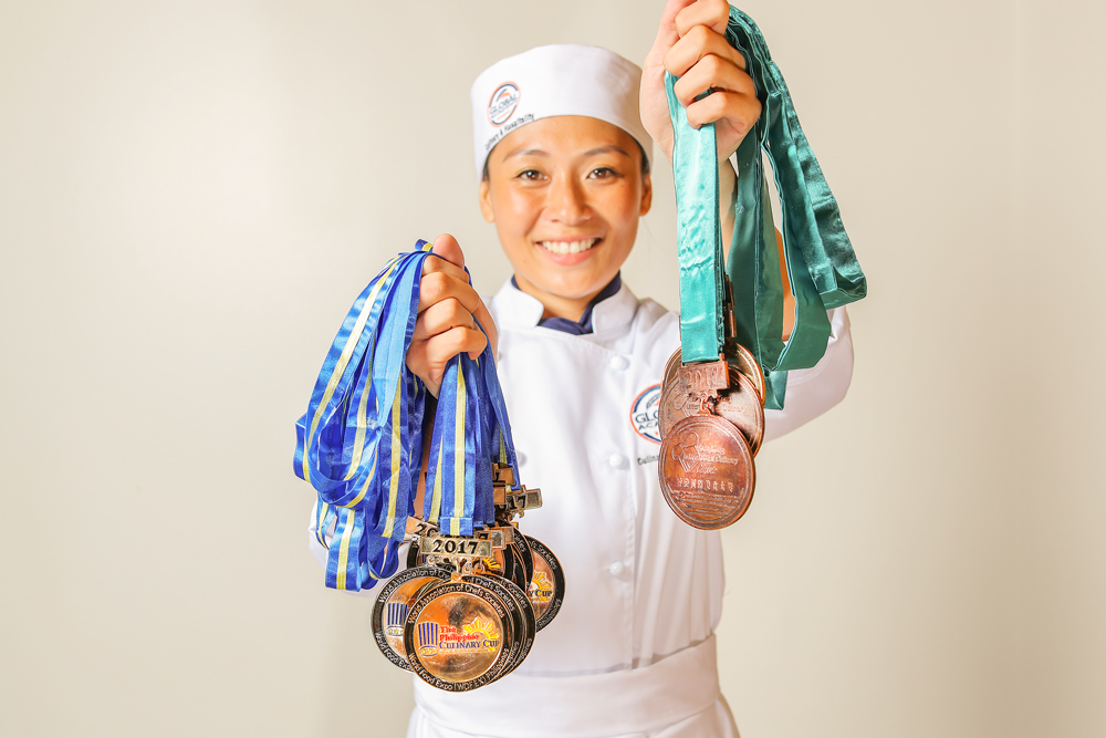 female chef smiling and holding up several medals in both hands