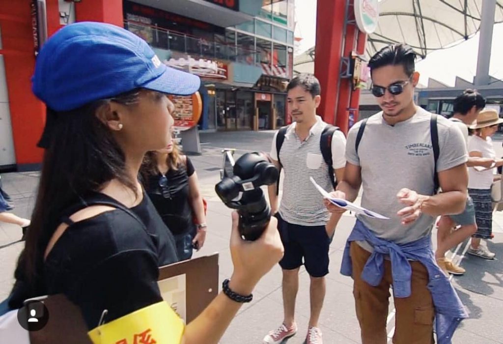 young woman holding a camera while facing two men, one of them reading a script