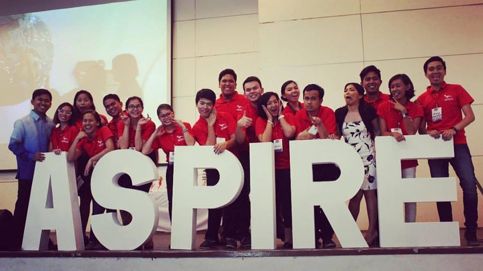 group of people in red t-shirts standing behind giant letters that spell "ASPIRE"