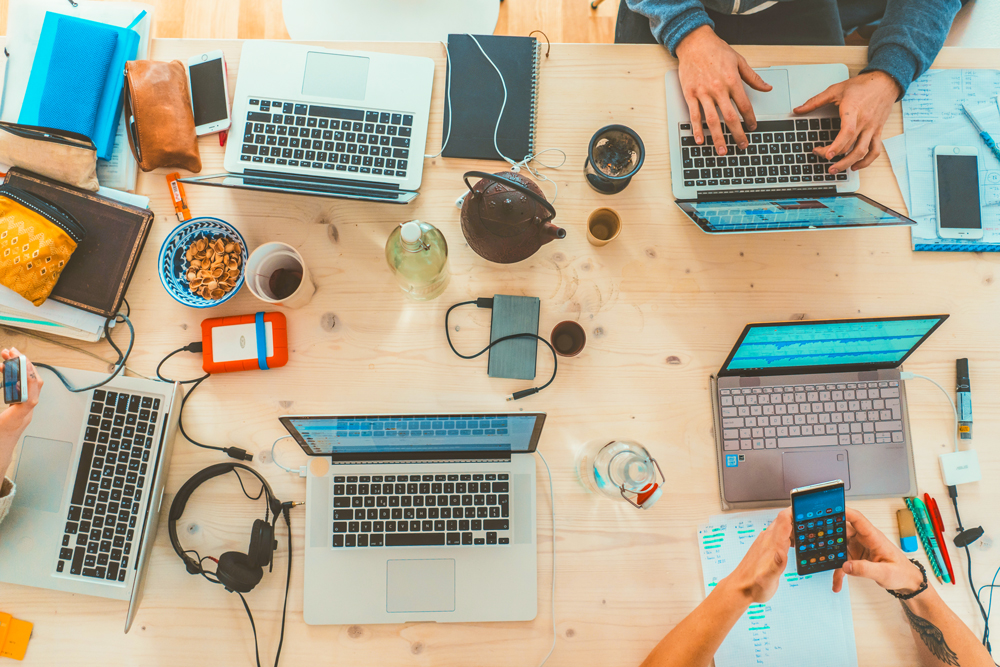 Overhead shot of hands working on laptops. Gadgets and headphones scattered across the table