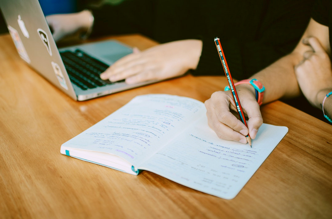 hand holding a laptop, another person's hand holding a pencil and writing on a notebook