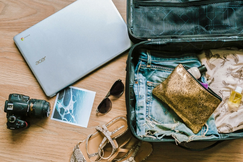 open luggage filled with clothes and a pouch. Laptop, shades, camera, and sandals on the floor