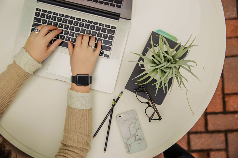 typing on silver laptop beside plant, notebook, glasses and phone