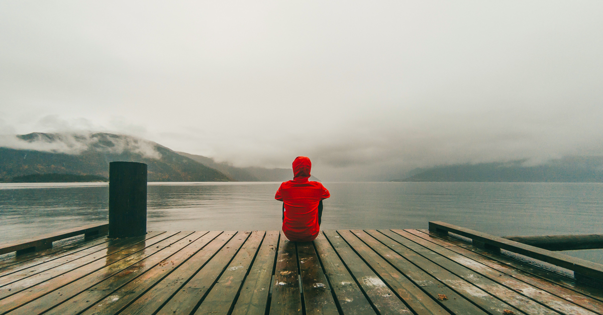 person wearing red hooded jacket sitting on brown and black wooden sea dock near mountains under gray sky during daytime