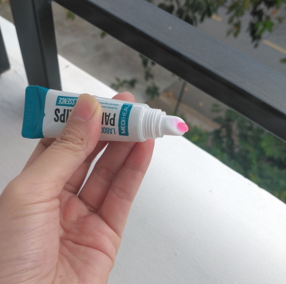 Cô gái cầm tuýp son dưỡng môi Mediheal màu xanh, sản phẩm giúp dưỡng ẩm và trị thâm môi, với giá chỉ 79k. Bài viết chia sẻ review mỹ phẩm và tìm kiếm son dưỡng loại nào tốt cho môi khô do ngủ máy lạnh.
