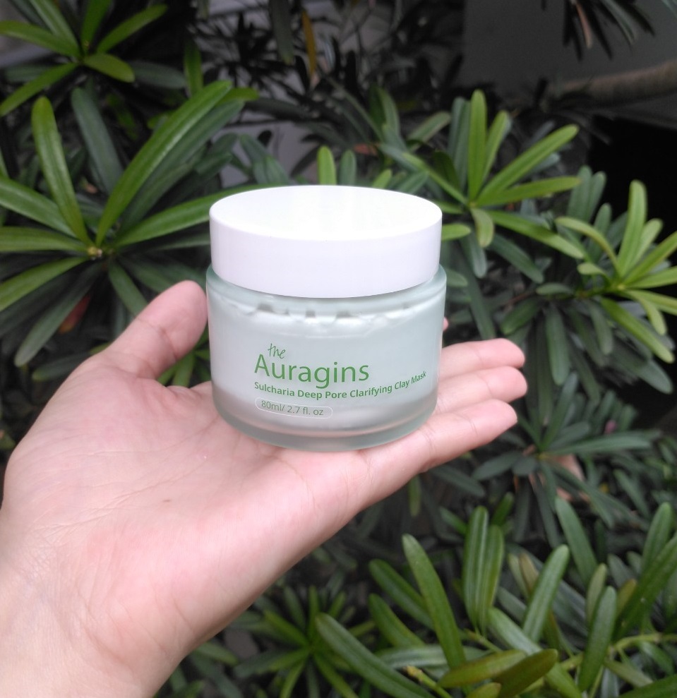 Mặt nạ đất sét The Auragins Sulcharia Deep Pore Clarifying Clay Mask được cầm trên tay, bao bì trong suốt, màu xanh dịu dàng giữa khung cảnh nhiều cây xanh. Mời bạn xem review mỹ phẩm, đặc biệt cho da dầu mụn.