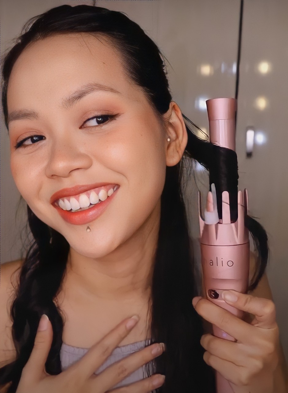 Mỹ Linh cầm máy uốn tóc tự động Halio, cười tươi và tạo kiểu tóc, chia sẻ về máy uốn tóc Halio trong video review. Khám phá ưu điểm của máy uốn tóc tự động, sự dễ sử dụng và hiệu quả trong việc tạo kiểu tóc bồng bềnh mà không làm tóc khô xơ.
