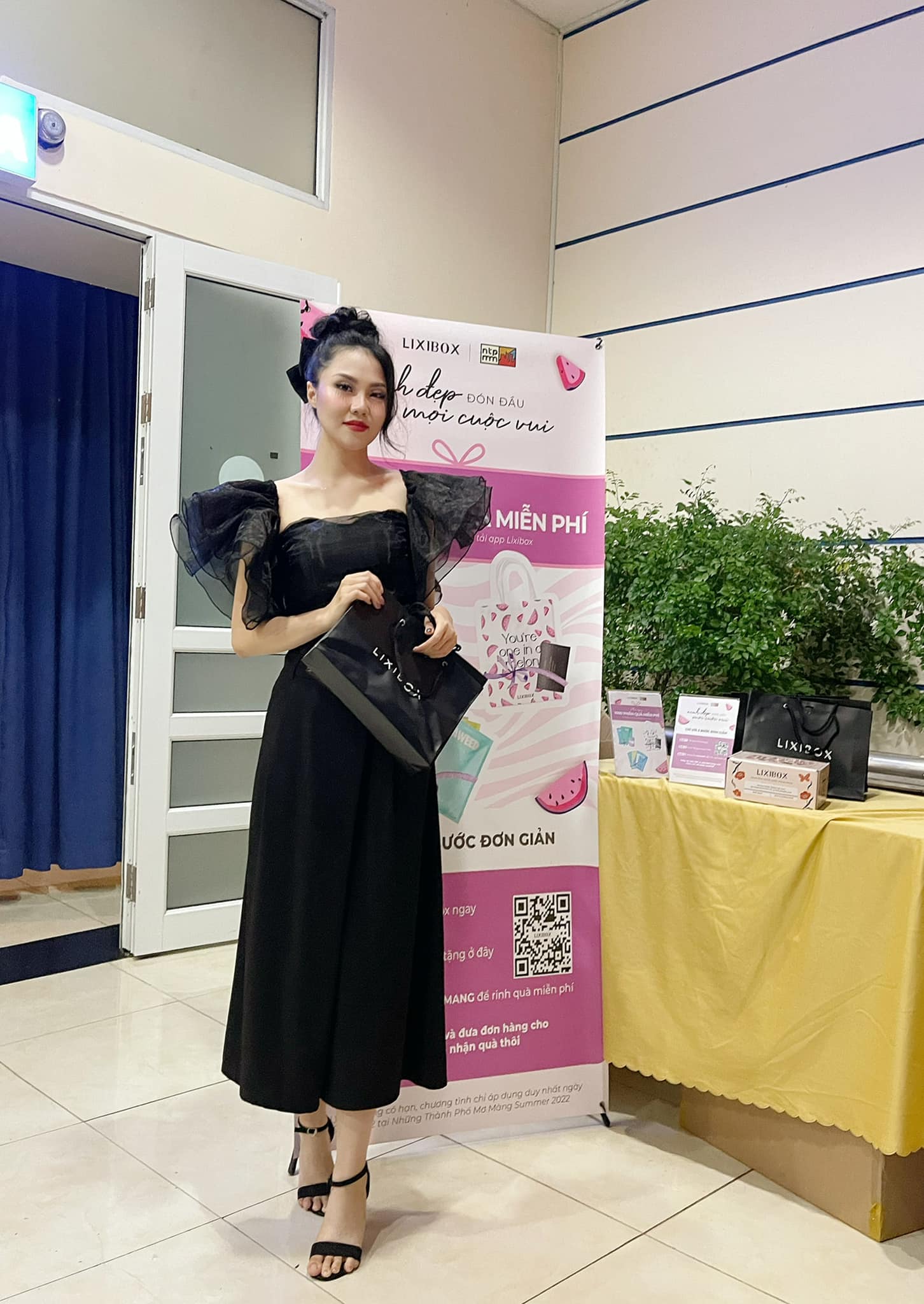 Cô gái trong trang phục đen đứng bên quầy quà tặng Lixibox, cùng với backdrop chương trình âm nhạc indie "Những Thành Phố Mơ Màng". Sự kiện diễn ra tại Nhà thi đấu Nguyễn Du, thu hút hơn 4000 khán giả, cùng với nhiều món quà hấp dẫn.