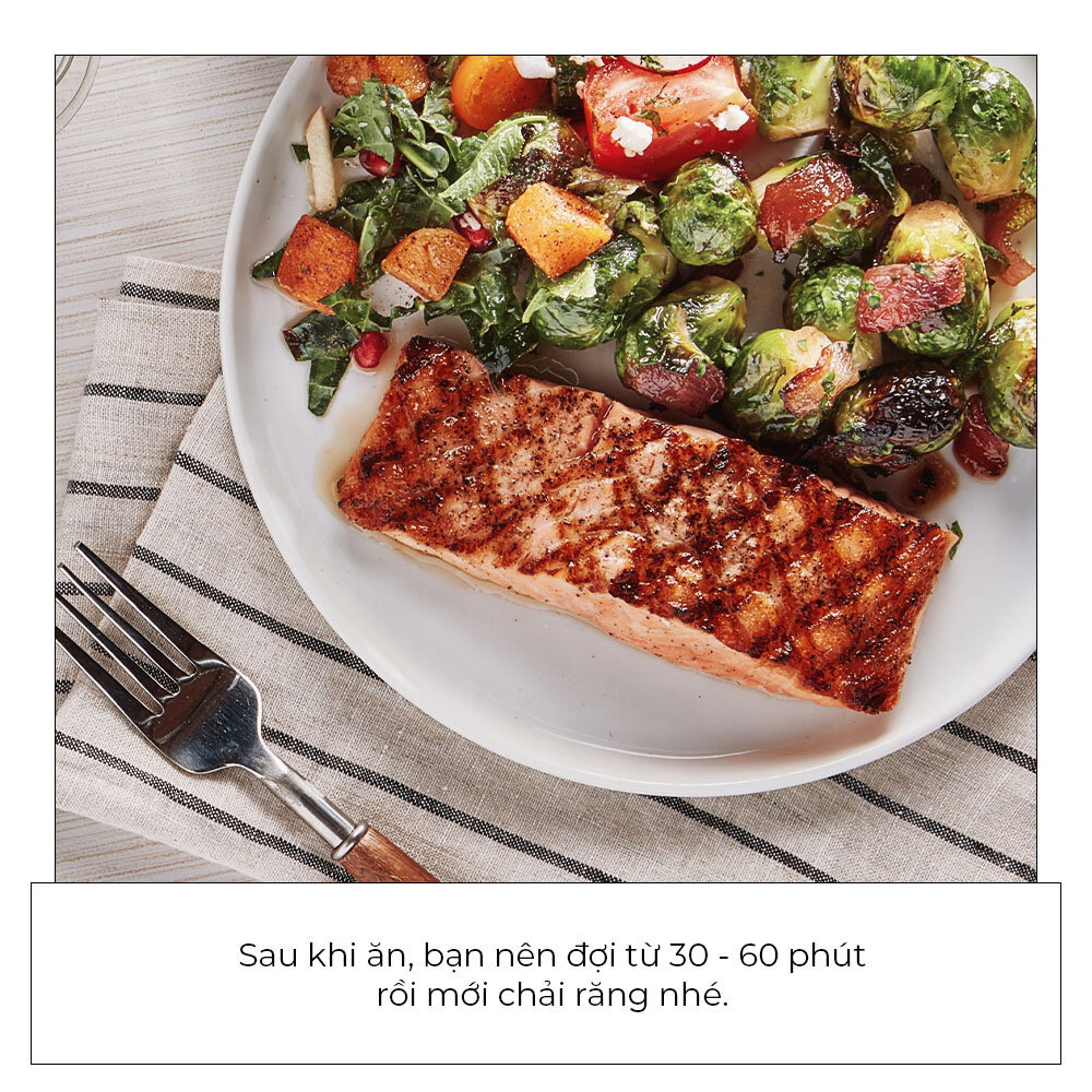 Bữa ăn với cá hồi nướng và salad, bên dưới có ghi chú khuyến nghị đợi từ 30 - 60 phút sau khi ăn mới nên chải răng. Thông điệp nhấn mạnh tầm quan trọng của thời gian trong chăm sóc răng miệng.