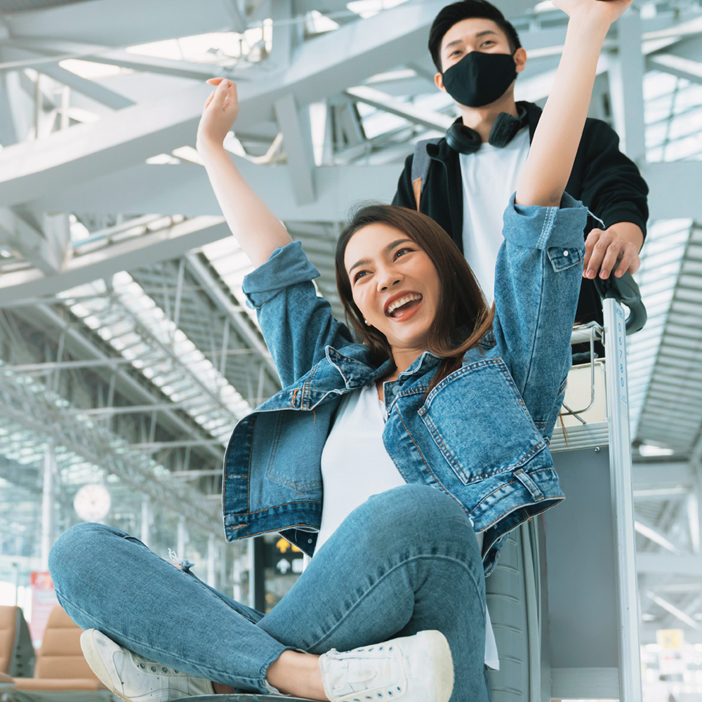 Khung cảnh hạnh phúc tại sân bay, một cô gái tươi vui ngồi trên xe hành lý, hai tay giơ cao thể hiện niềm phấn khích. Hình ảnh thể hiện sức mạnh của sự kết nối trong tình yêu xa và bí quyết yêu xa bền vững.