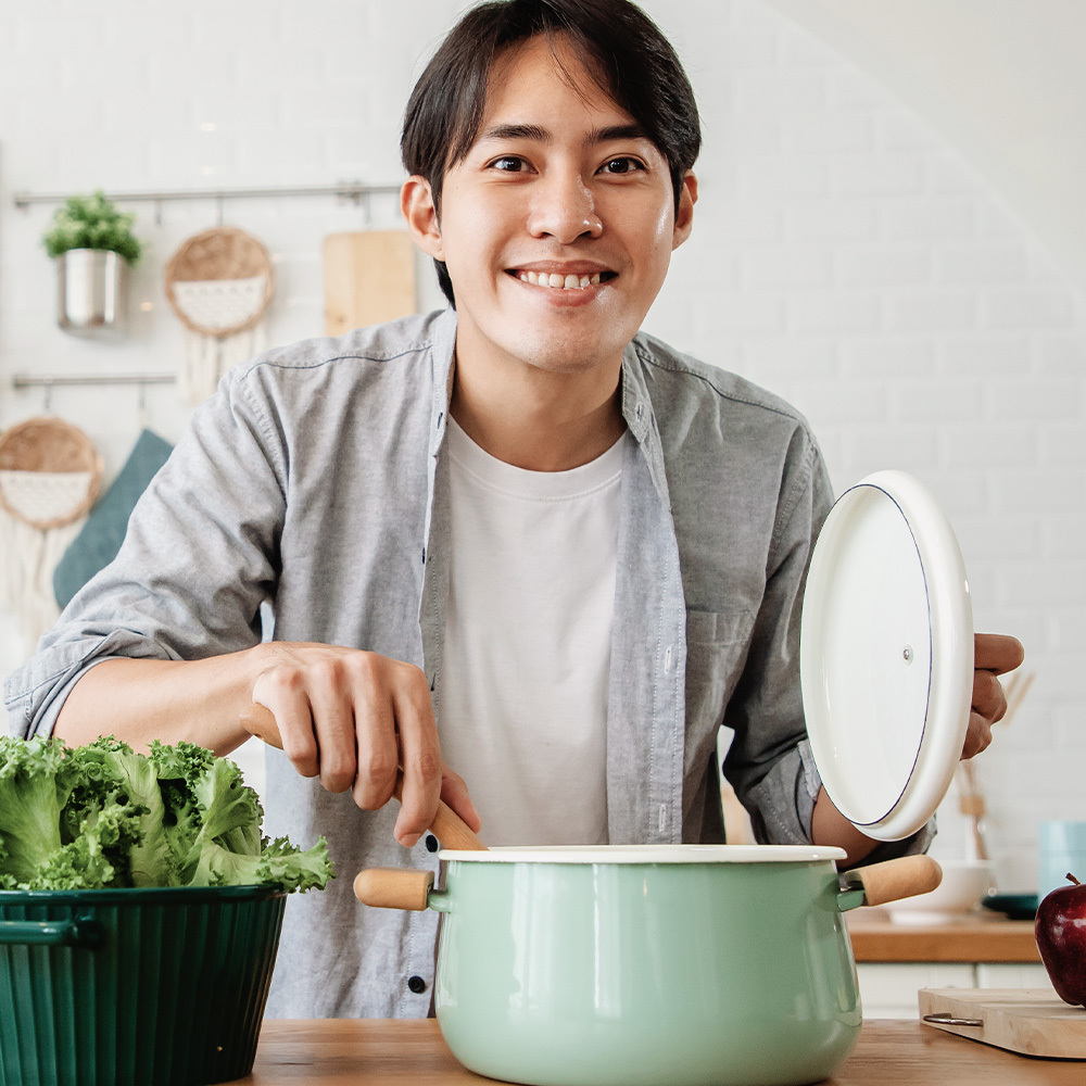 Một người đàn ông đang nấu ăn trong bếp, mỉm cười và mở nắp nồi, xung quanh có rau xanh. Bài viết cung cấp bí quyết yêu xa bền vững cho những cặp đôi yêu nhau, bao gồm các hoạt động như nấu ăn cùng nhau để giữ lửa tình cảm.