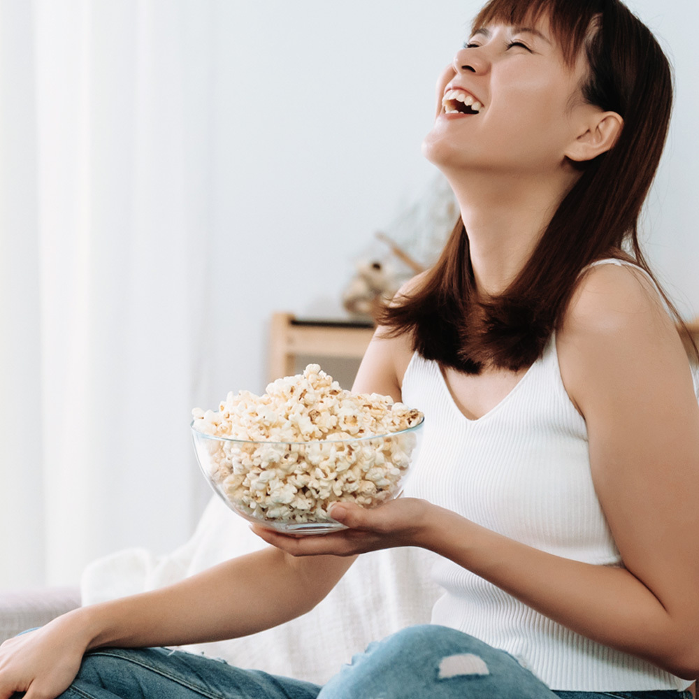 Cô gái cười tươi, cầm bát bỏng ngô trong không gian thoải mái. Bài viết chia sẻ bí quyết yêu xa và 5 hoạt động nên làm để giữ gìn mối quan hệ bền lâu, khuyến khích sử dụng bàn chải điện Halio trong chăm sóc sức khỏe răng miệng.