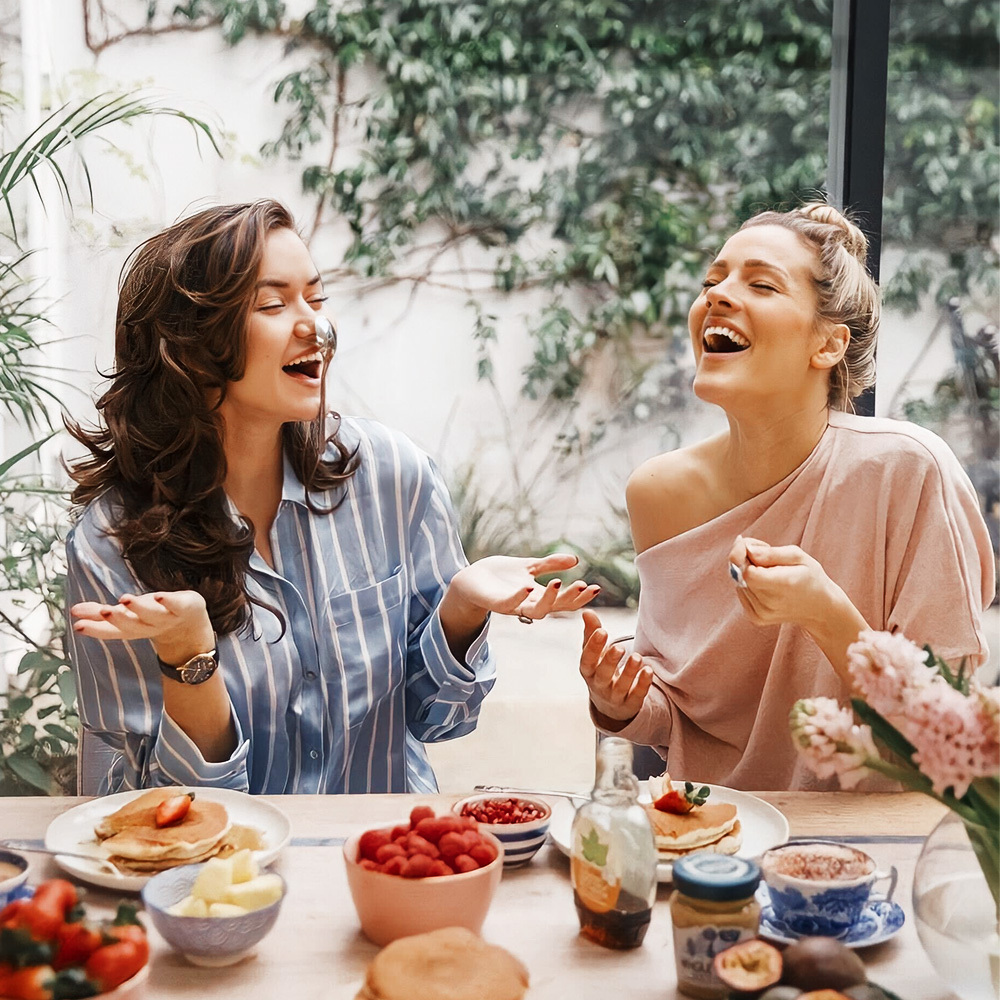 Hai phụ nữ trẻ vui vẻ bên bàn ăn sáng, chia sẻ tiếng cười và niềm hạnh phúc trong ngày Quốc tế Hạnh phúc 20 tháng 3, thể hiện ý nghĩa của 6 điều nhất định phải làm trong ngày này.