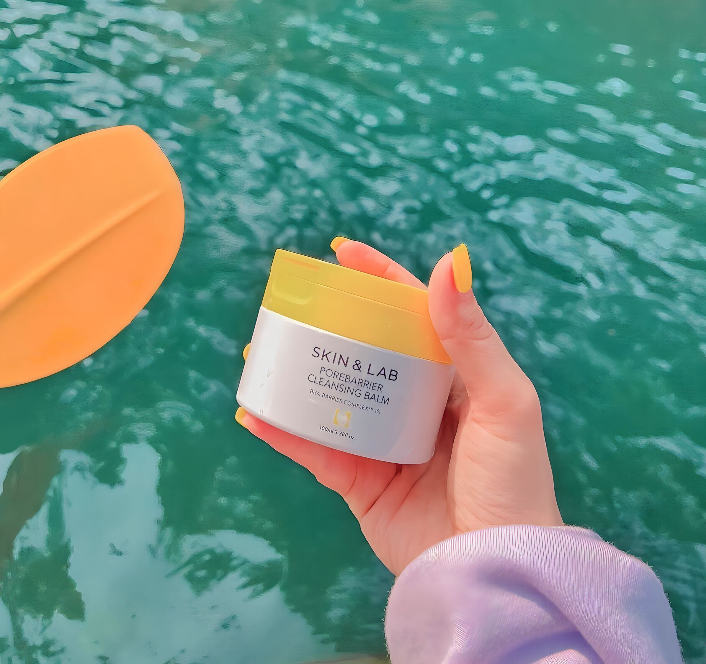 Sáp tẩy trang SKIN&LAB được cầm trên tay, với bao bì màu trắng và vàng nổi bật, giữa khung cảnh nước xanh, mang lại cảm giác tươi mới. Đọc bài Lixibox Review để khám phá hiệu quả thực sự của sản phẩm sáp tẩy trang này.