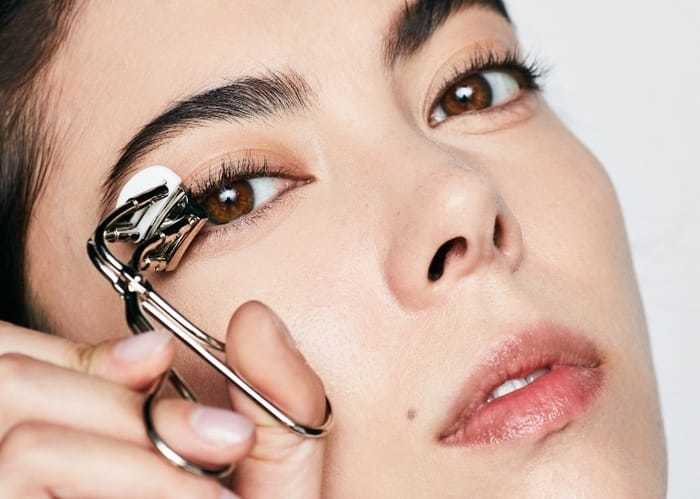 Cô gái trẻ đang sử dụng dụng cụ uốn mi, thể hiện cách trang điểm cho người lớn tuổi. Bài viết chia sẻ 10 mẹo trang điểm cực đơn giản giúp gương mặt trở nên trẻ trung hơn, phù hợp với Hot Trend trang điểm trẻ hơn tuổi.