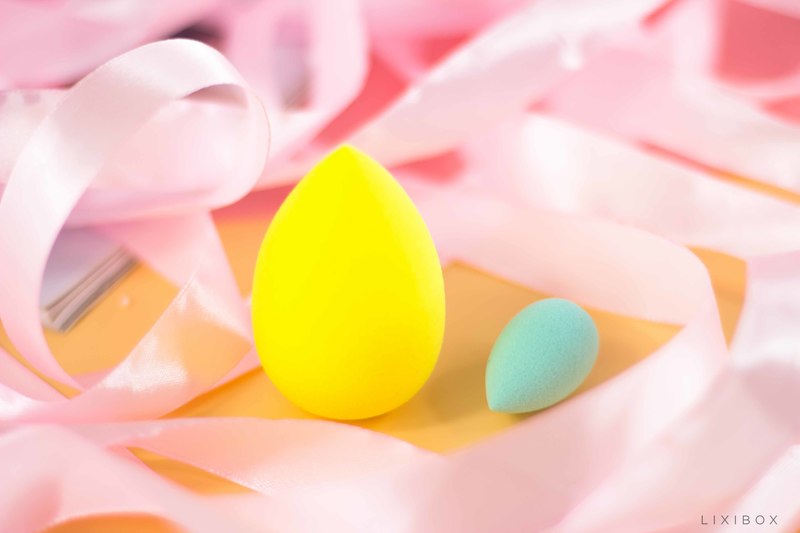Hai miếng bông mút làm đẹp với màu sắc tươi sáng, nổi bật trên phông nền trang trí pastel, thể hiện xu hướng làm đẹp hot trend. Bông mút trang điểm hình trứng lớn màu vàng và nhỏ màu xanh, tượng trưng cho các sản phẩm hỗ trợ lớp nền hoàn hảo.
