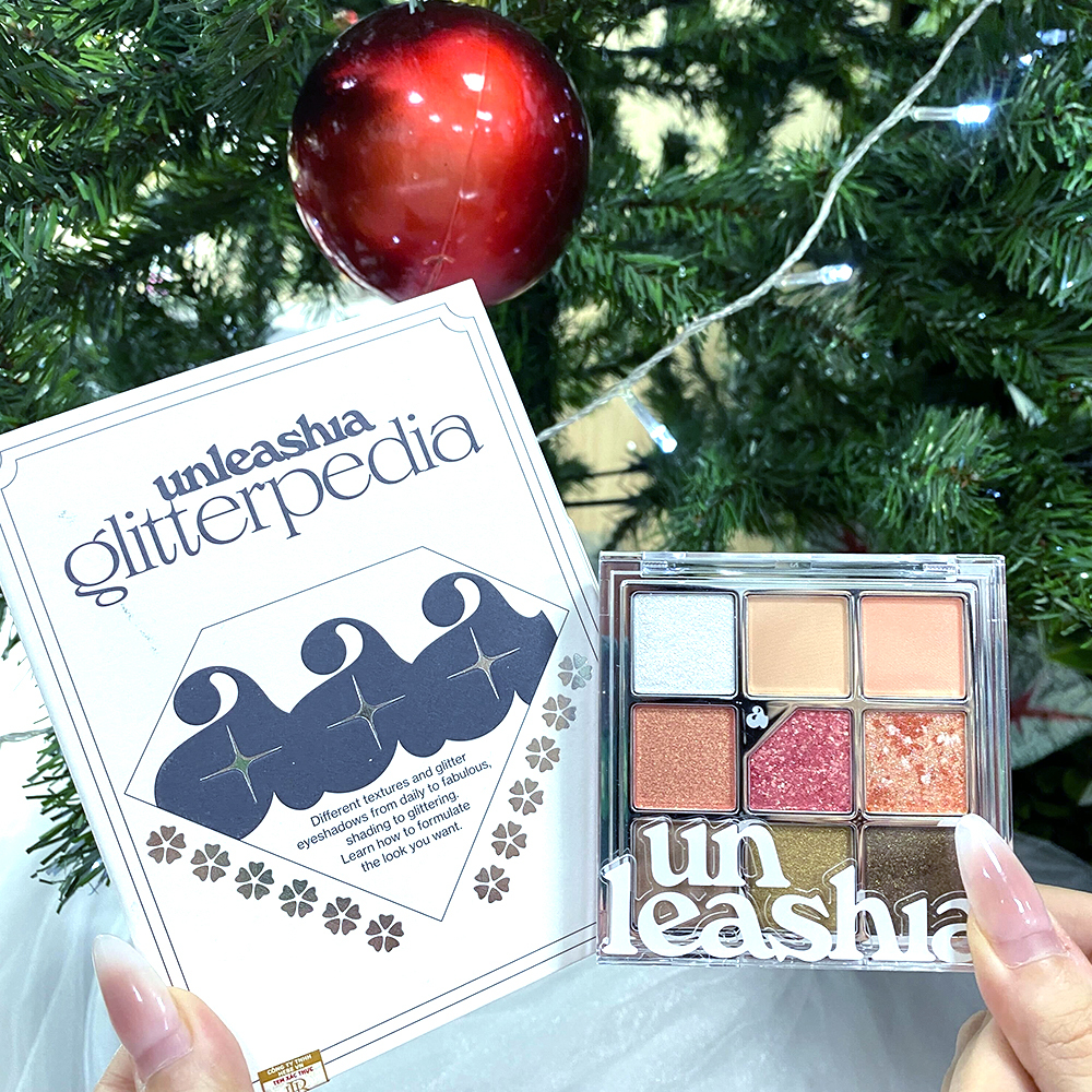 Hộp quà Lixibox với sản phẩm unleashia glitterpedia kết hợp giữa phấn mắt và nhũ trên nền cây thông Giáng sinh, thể hiện sự lôi cuốn và tiện lợi cho việc trang điểm mùa lễ. GWP Mua Lành giúp chữa lành bản thân hậu tan vỡ.