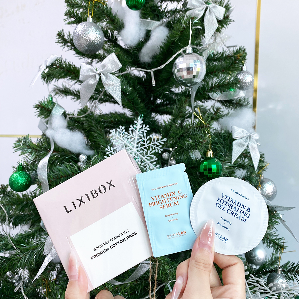 Hộp quà GWP từ Lixibox với sản phẩm dưỡng da, bao gồm bông tẩy trang 3 trong 1, serum Vitamin C sáng da và kem dưỡng ẩm Vitamin B, được đặt dưới cây thông Noel, thể hiện sự săn sale cuối năm và tím kiếm sản phẩm chữa lành bản thân hậu tan vỡ.