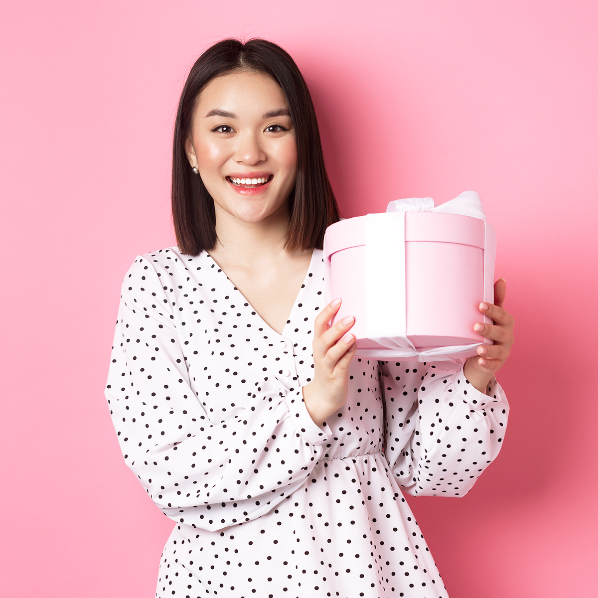 Người phụ nữ tươi cười cầm món quà trong tay, đứng trước nền hồng nhẹ nhàng, thể hiện niềm vui trong việc tặng quà. Bài viết cung cấp những tips hữu ích để hiểu sở thích quà tặng của phụ nữ, giúp các chàng trai dễ dàng lựa chọn quà phù hợp.