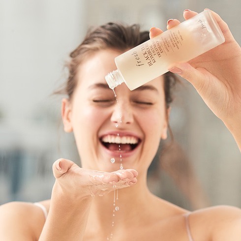 Người phụ nữ vui vẻ đang áp dụng sản phẩm chăm sóc da cơ bản, kiểm tra quy trình skin care routine của mình. Bài viết nhấn mạnh tầm quan trọng của việc hiểu rõ các bước chăm sóc da và cách sắp xếp đúng đắn để đạt hiệu quả tốt nhất.