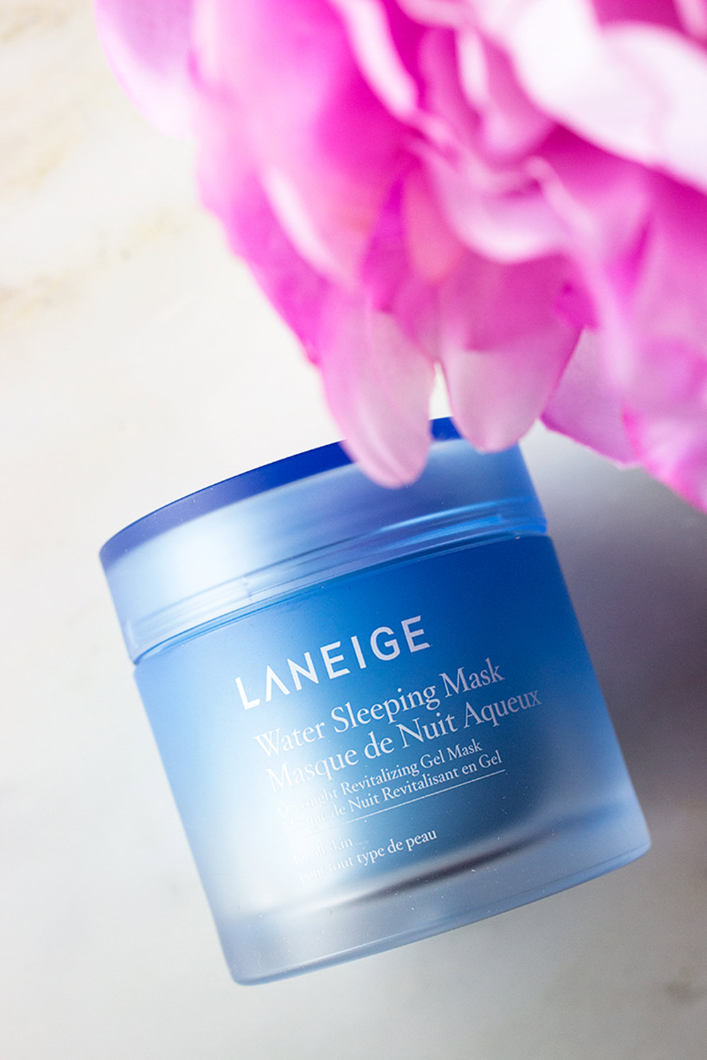 Hộp mặt nạ ngủ Laneige Water Sleeping Mask với màu xanh dương, nằm trên bề mặt trắng, bên cạnh hoa hồng hồng, là sản phẩm lý tưởng cho chăm sóc da, giúp cải thiện độ ẩm và phục hồi làn da trong mùa Sài Gòn ẩm ương.