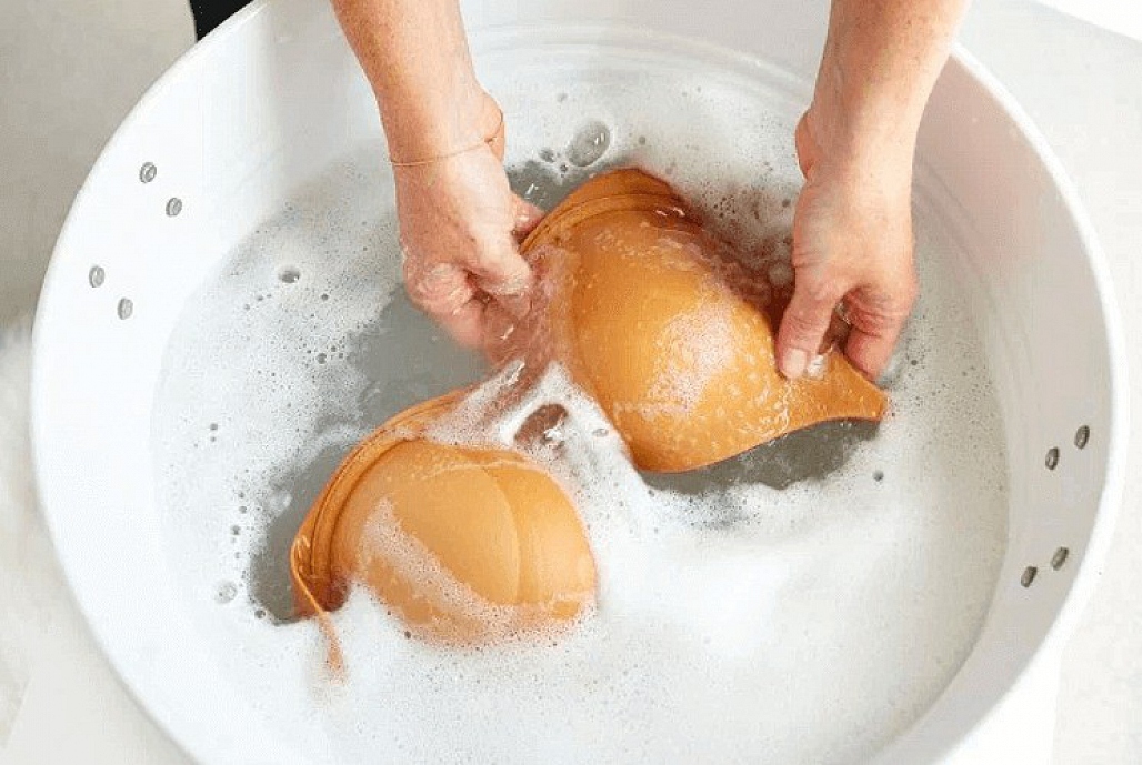 Cách bảo quản nội y đúng cách để tránh vi khuẩn, hình ảnh người đang giặt áo ngực màu da trong chậu nước xà phòng. Điều này rất quan trọng để bảo vệ sức khỏe và duy trì độ bền của bikini.