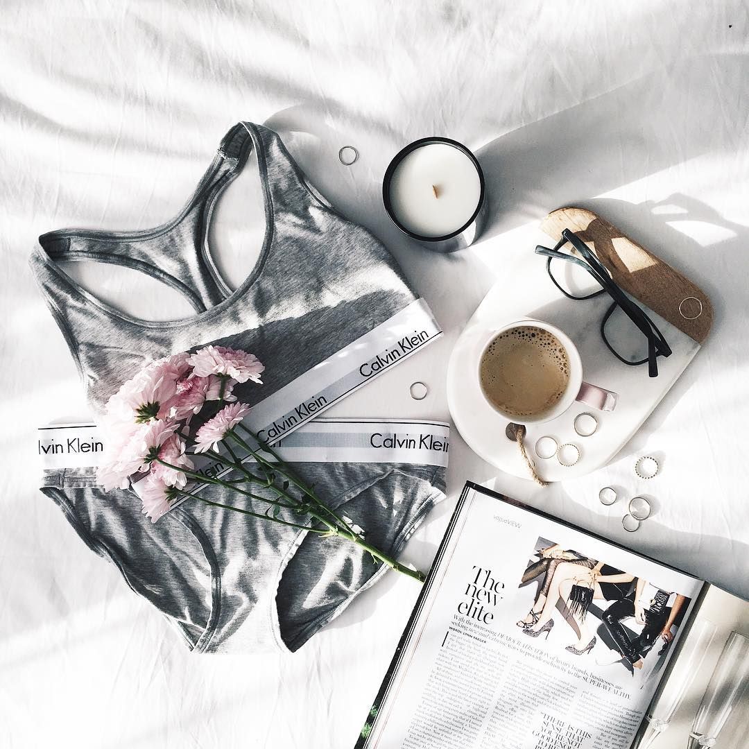 Bộ nội y Calvin Klein màu xám nằm trên nền vải trắng, bên cạnh là tạp chí thời trang, cốc trà, nhánh hoa và một số phụ kiện lấp lánh. Nội y thể hiện nét cá tính độc đáo của phái đẹp, tạo dấu ấn riêng biệt.
