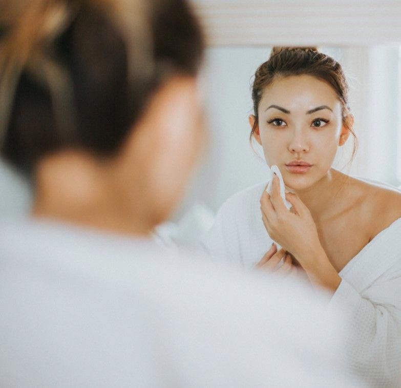 Người phụ nữ trẻ tuổi chăm sóc da mặt trong gương, thể hiện sự quan tâm đến quy trình dưỡng da. Bài viết hướng dẫn quy trình dưỡng da theo từng độ tuổi và các sản phẩm chăm sóc da cơ bản.