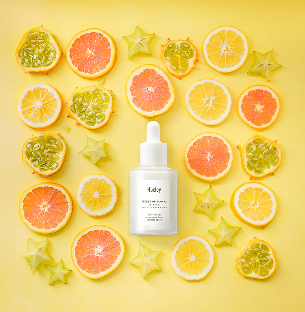 Hình ảnh serum Huxley "Secret of Sahara" giữa các loại trái cây tươi như chanh, bưởi, và sao, nhấn mạnh trong quy trình dưỡng da cơ bản với Halio Ion để cải thiện hiệu quả dưỡng trắng và trị thâm.