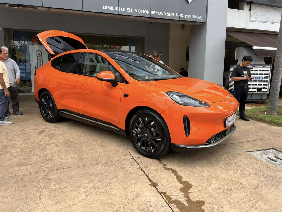 Kereta elektrik GAC Aion Hyptec HT berwarna oren terang dengan rim aloi hitam dipamerkan di luar bilik pameran. Pintu gullwing kereta terbuka, menonjolkan reka bentuknya yang aerodinamik dan bergaya. Beberapa orang berdiri berdekatan, memerhatikan kenderaan tersebut