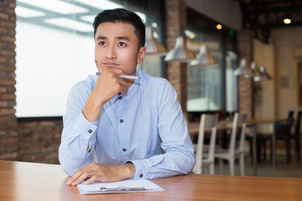 Young man in blue shirt sitting at table with clipboard, thinking deeply and holding pen near chin.