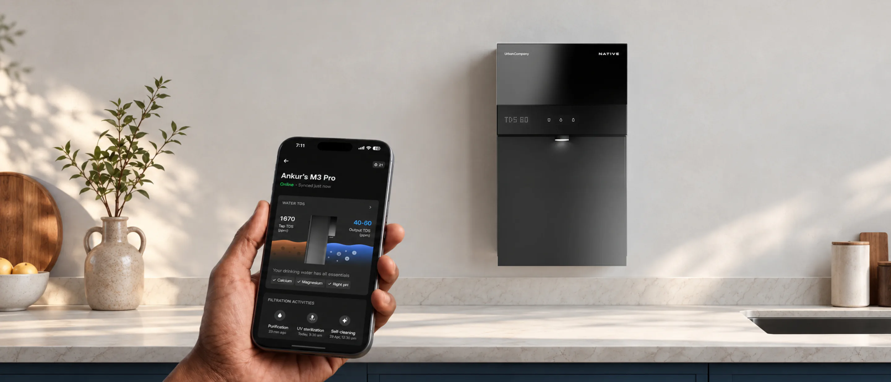 Hand holding a phone with the Native M3 Pro open in the UC app, the purifier mounted on the kitchen wall behind.