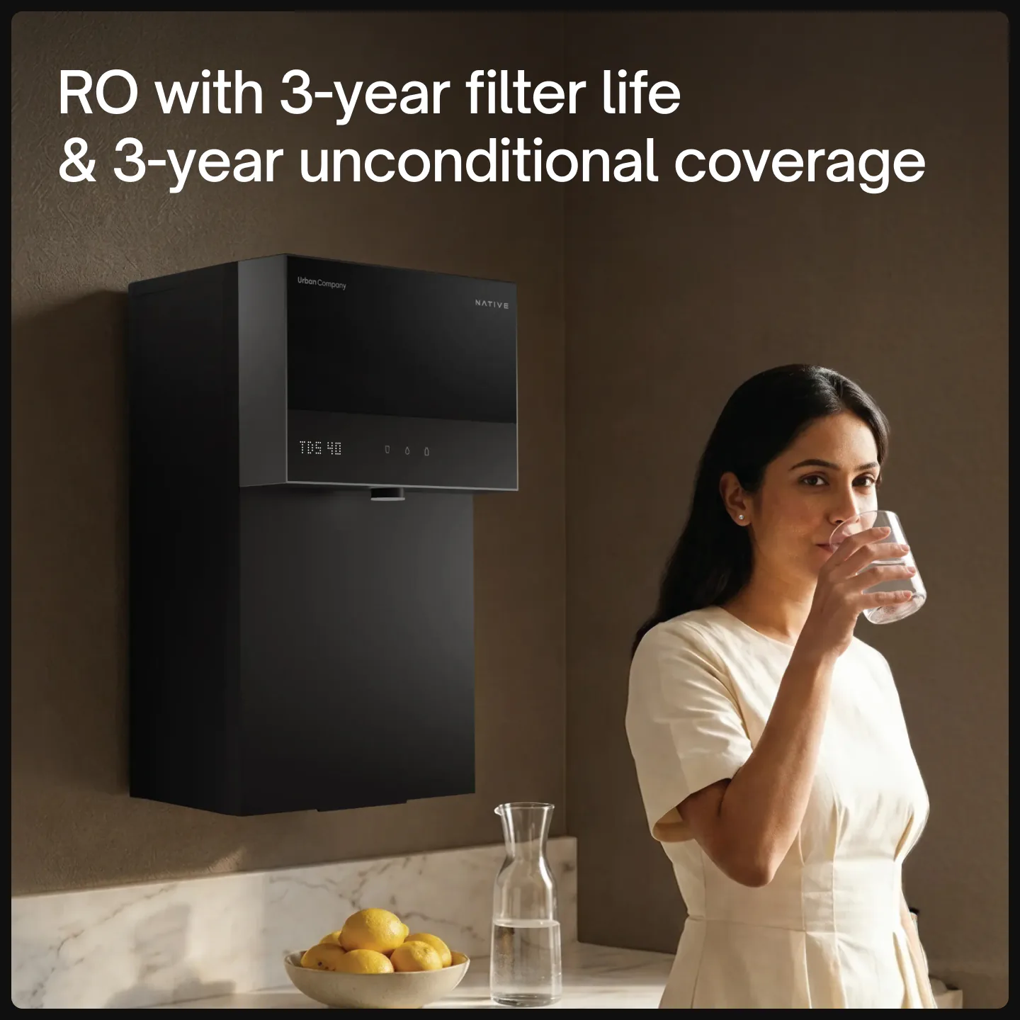 Woman drinking a glass of water next to a wall-mounted Native M3 Pro RO purifier in a kitchen. Text: RO with 3-year filter life & 3-year unconditional coverage.