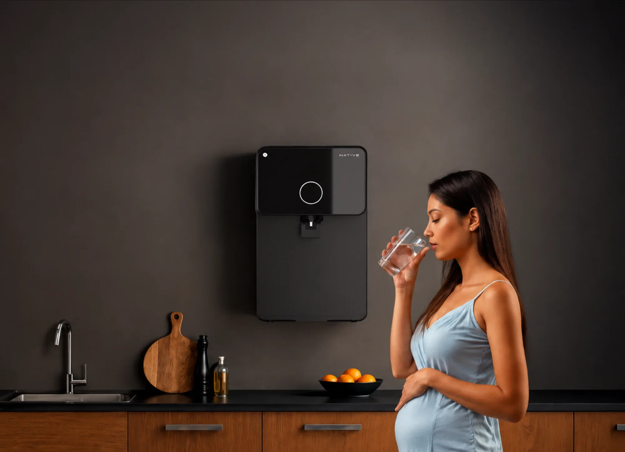A pregnant woman drinks a glass of water beside a wall-mounted Native M1 in a dark kitchen.